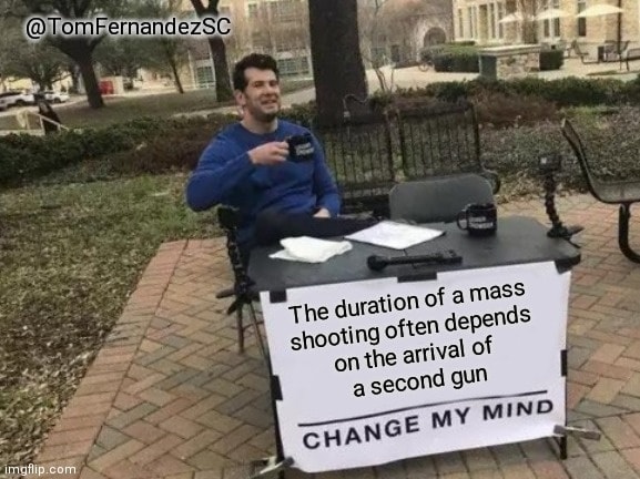 A person sitting at a table outdoors with a sign that says, "The duration of a mass shooting often depends on the arrival of a second gun. Change my mind." They are holding a mug and appear to be inviting discussion.