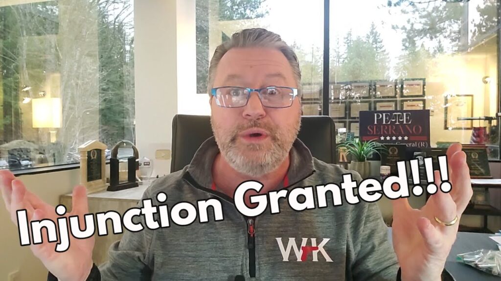 A man with glasses and a beard is sitting in an office with large windows. He has his hands raised and is looking surprised. Text over the image reads, "Injunction Granted!!!" The background shows trees and office decor.