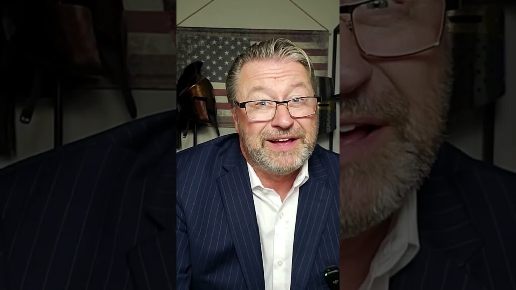 A bearded man wearing glasses and a pinstripe suit jacket smiles while sitting indoors. An American flag and cowboy hats can be seen in the background.