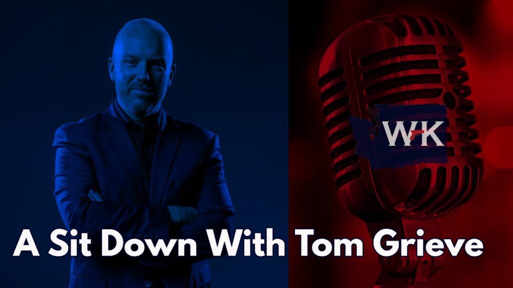 A person in a suit stands with folded arms in blue lighting on the left. A red microphone is on the right. The text reads "A Sit Down With Tom Grieve" at the bottom.