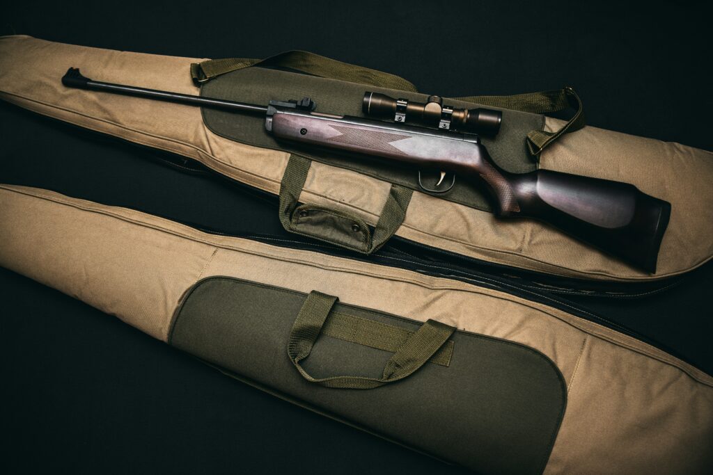 A rifle with a scope rests on top of two padded gun cases, one tan and one olive green, against a dark background. The cases have handles and pockets, and the rifle appears polished and well-maintained.