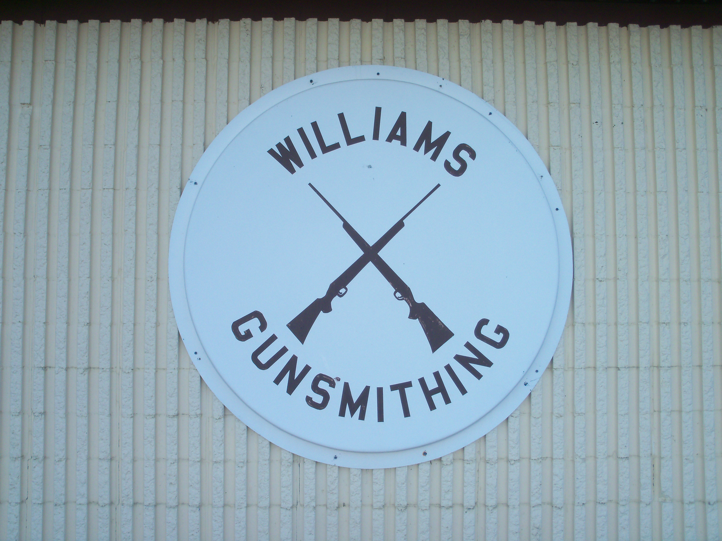All Listings 2 A white circular sign on a ribbed wall features two crossed rifles in the center. The text "Williams Gunsmithing" arches above and below the rifles in bold, black letters.
