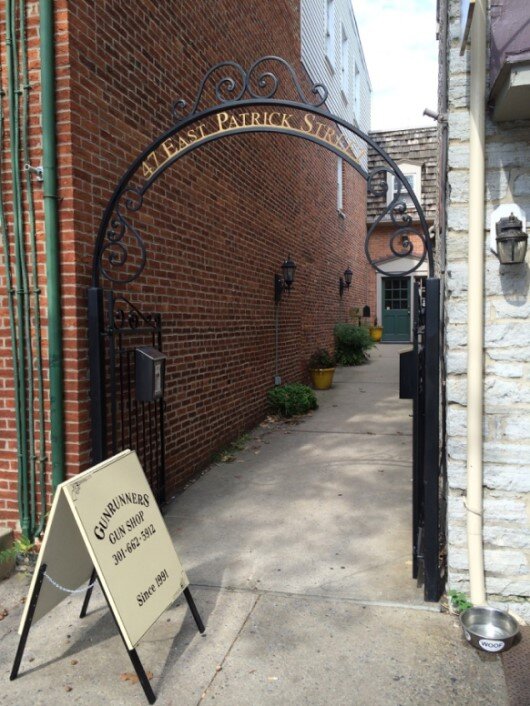 Search Result 1 A wrought iron archway leads to a narrow alley lined with brick walls. A sign beside the alley reads "Gunrunners Gun Shop" with a phone number, and there are yellow potted plants along the path.