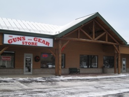 A wooden storefront with a large sign reading "The Guns and Gear Store" above the entrance; snow is on the ground in front of the building.