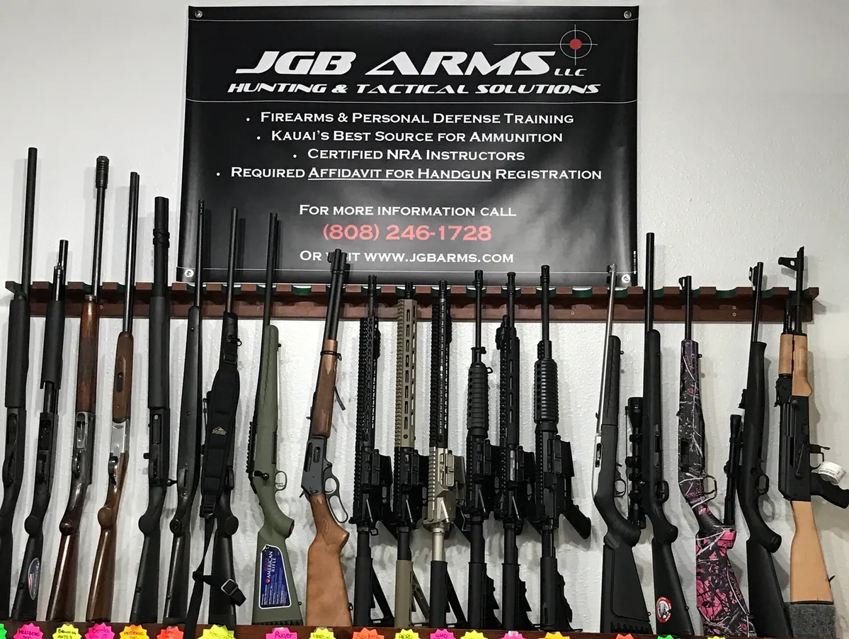 Hawaii Gun Clubs 4 A display of various rifles and shotguns lined up against a wall beneath a banner. The banner reads: "JGB ARMS LLC Hunting & Tactical Solutions." Contact details and additional services are listed on the banner.