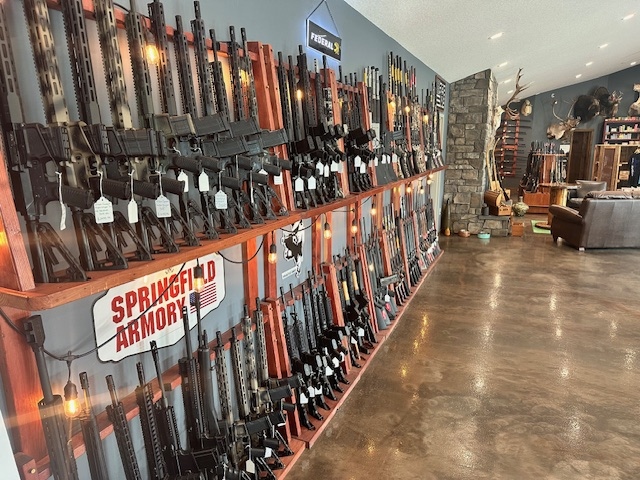 Rows of rifles are displayed on wooden racks along the wall of a gun store, with signs for Springfield Armory and other brands visible. The store has polished concrete floors and mounted animal heads on the far wall.