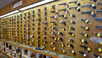 A wooden wall display in a store shows dozens of handguns, each with a yellow price tag. Below the guns, shelves hold boxes and accessories. The display stretches across the length of the wall.