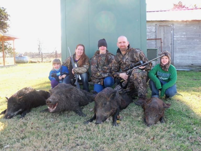 Single Category 2 A group of people wearing camouflage clothing are posed kneeling on grass. They are holding rifles and are surrounded by several wild boars lying on the ground. A green structure and a wooden building are in the background.