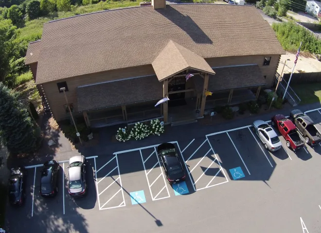 All Listings 5 Aerial view of a brown building with a pitched roof, surrounded by trees. In front, a parking lot with several cars and marked accessible parking spaces. Flags are displayed near the entrance. Shadows cast on the pavement indicate a sunny day.