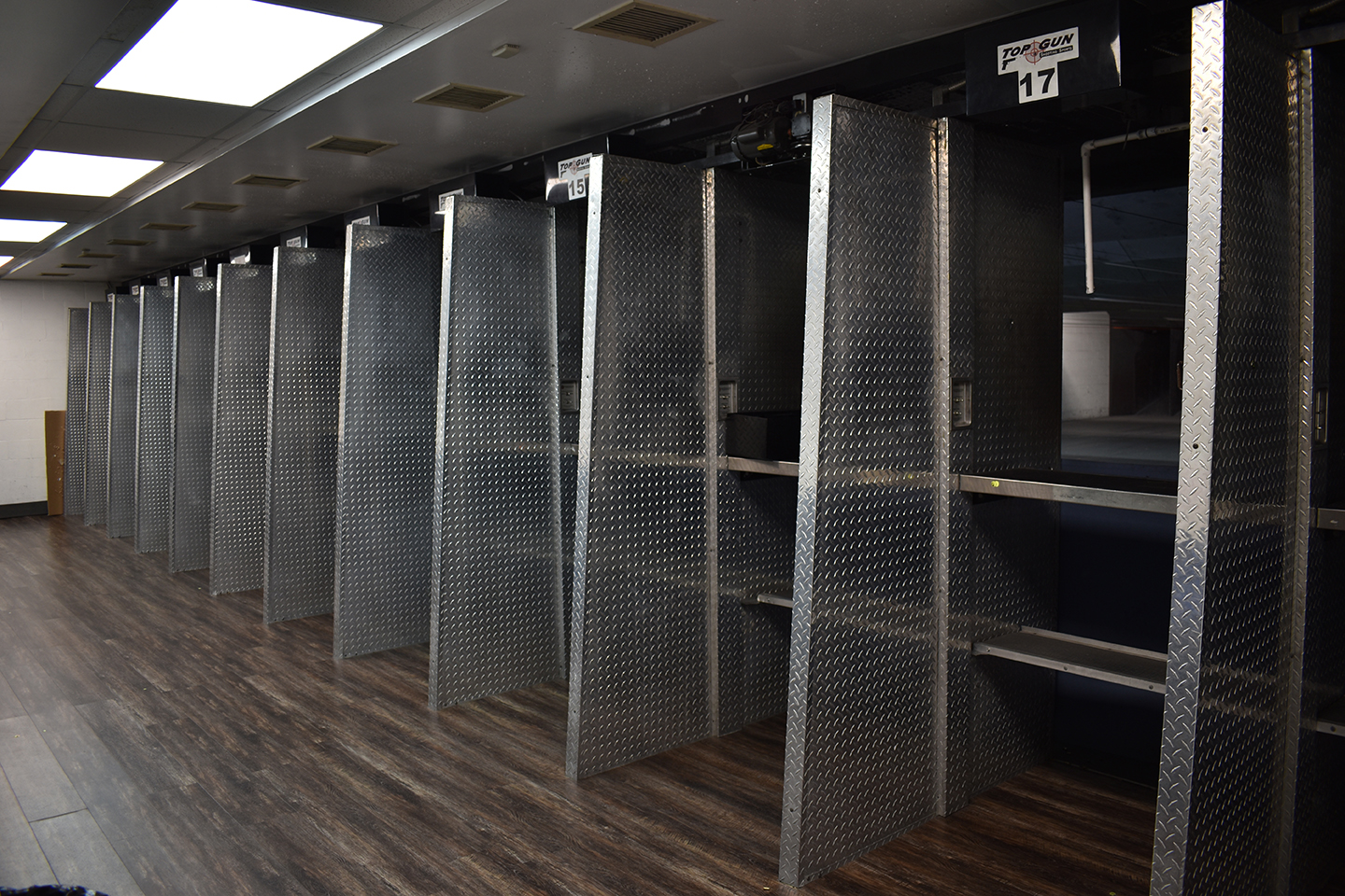Missouri Gun Clubs 4 A row of empty shooting range stalls with metallic dividers, numbered signs above each booth, and dark wood flooring. The stalls face towards a back wall with targets not visible in the image.