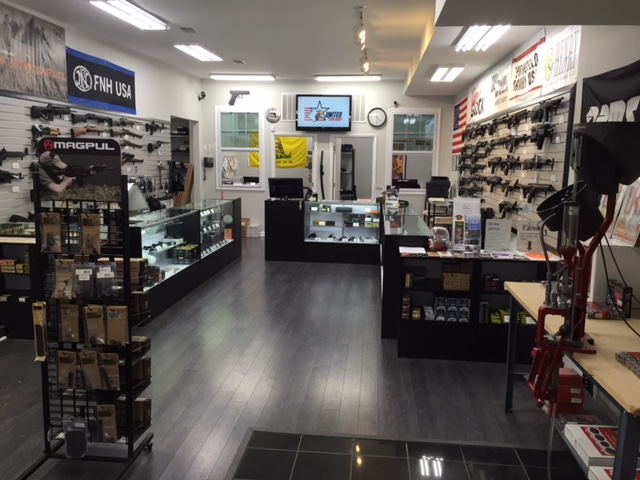Search Result 3 Interior of a gun store featuring display cases with firearms on the walls. The room is brightly lit with wooden flooring, and there are various accessories and products on shelves and racks, along with a counter and a television screen.