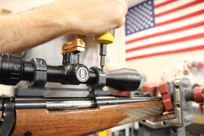 Single Category 4 Person working on a rifle scope alignment with tools, inside a workshop. An American flag is visible in the background.