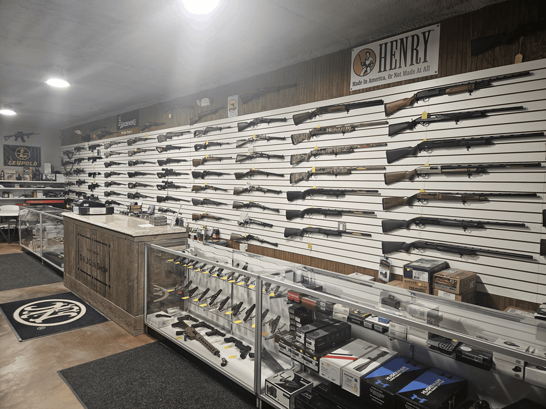 A gun store interior with rifles displayed on wall racks, glass display cases filled with handguns and accessories, and signs for Henry and Leupold brands visible above and on the counter.
