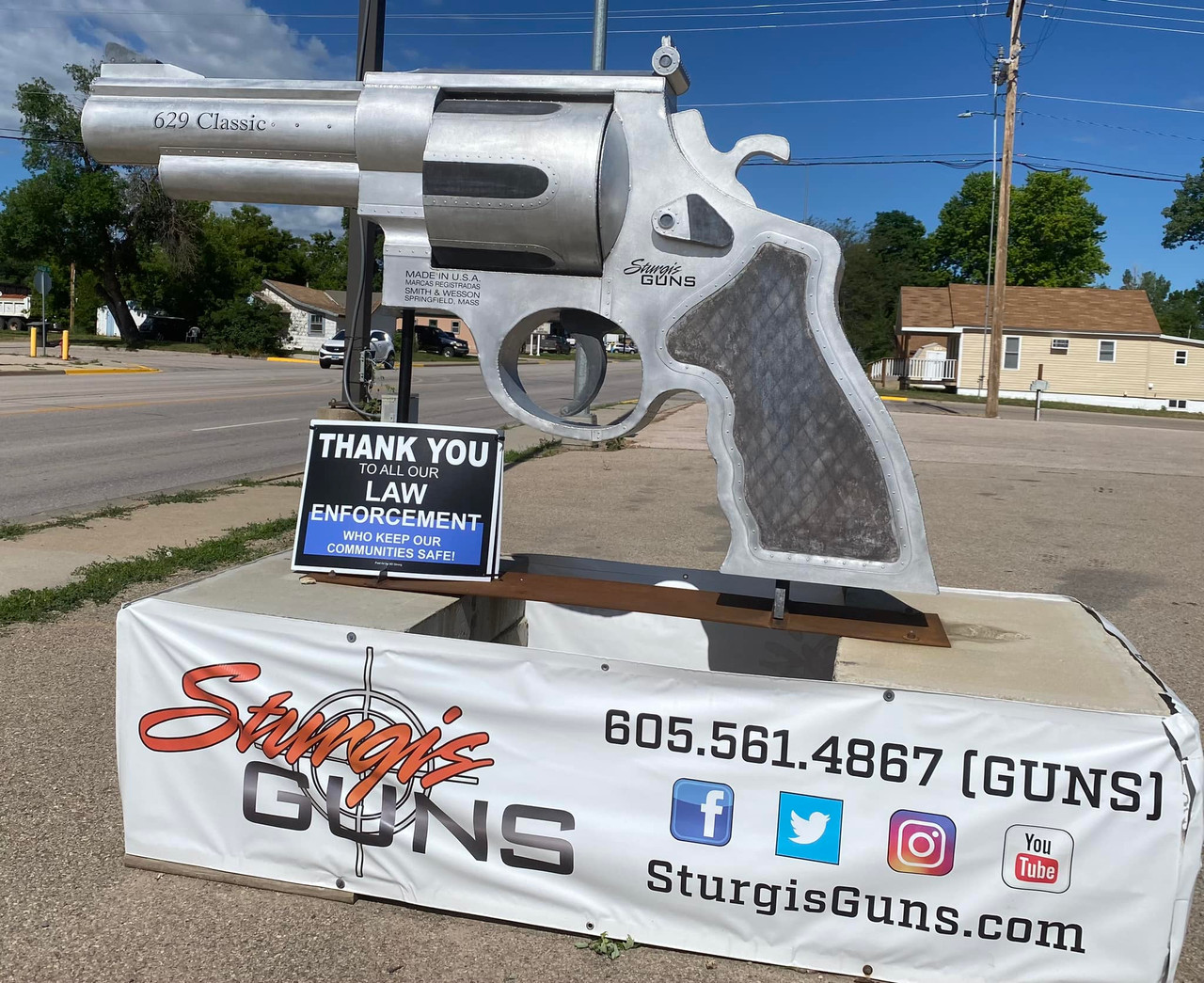 A large metal sculpture of a revolver is displayed on a street corner. A sign thanking law enforcement is in front, and a banner below advertises "Sturgis Guns" with contact info and social media icons.