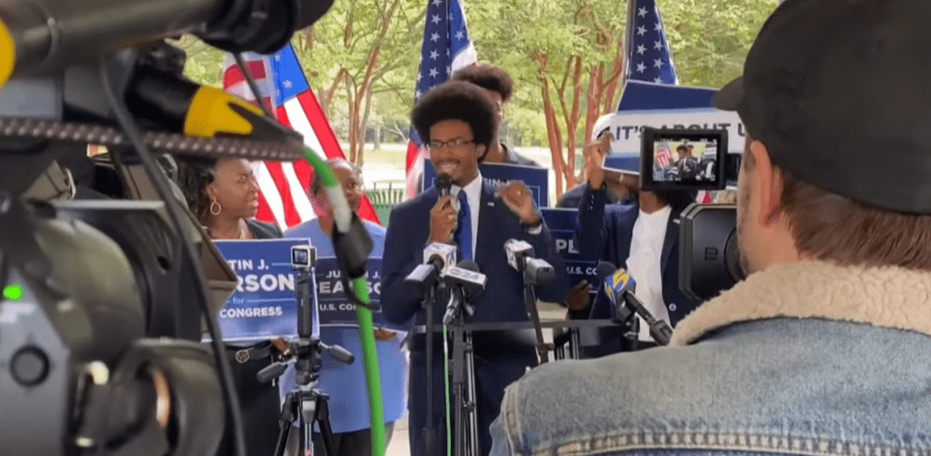 A man in a suit speaks at a podium with microphones during an outdoor press conference, surrounded by supporters holding campaign signs and American flags, while cameras and a person in a cap film the event.
